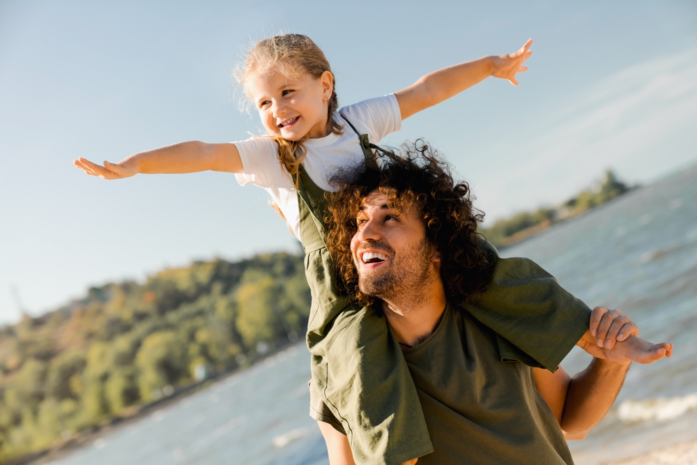 Father carrying his daughter on his shoulders at the lakeshore, both with arms outstretched